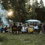 A group of adults relaxing around a campfire in a scenic outdoor setting with tents.