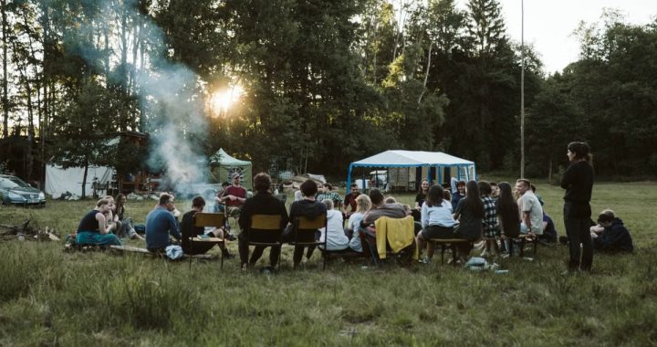 A group of adults relaxing around a campfire in a scenic outdoor setting with tents.